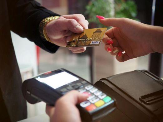 A hand handing a Visa card to another hand containing a credit card machine.